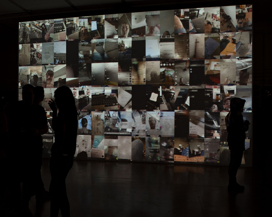 Three people standing in front of a wall-size projection of user_is_present (grid series), a gridded set of collaged video portraits