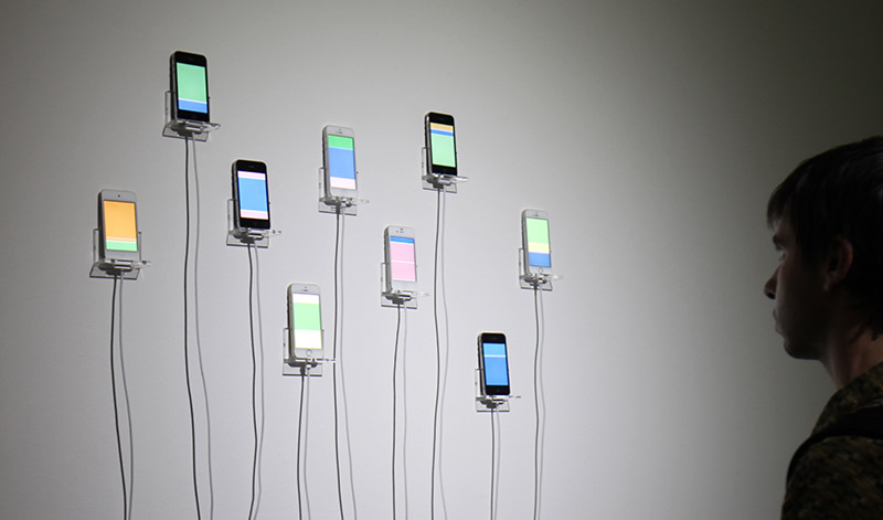 Man observes art installation with nine iPhones mounted on the wall showing blocks of bright colors on their screens, simulating the infinite scrolling motions of social media