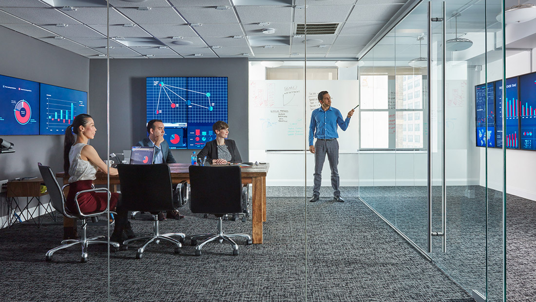 Four people collaborating in a high-tech Oblong Mezzanine conference room with many high-resolution screens on the walls, multiple laptops, and a gestural pointing device