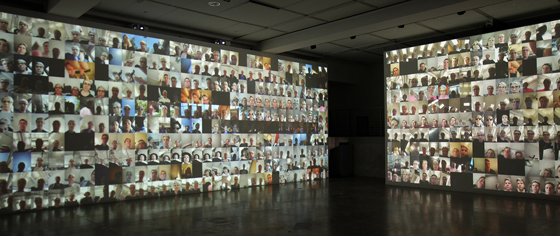 Two walls covered in a video projection of a grid of many smartphone selfie camera videos from the same user in phonelovesyoutoo: matrix