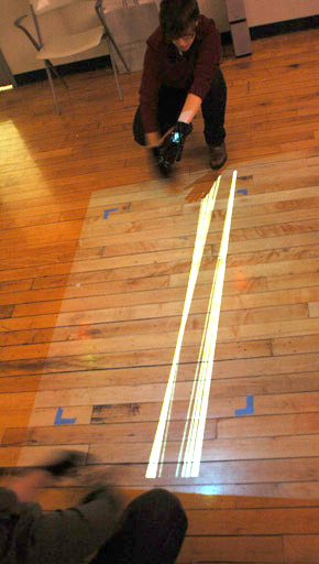 Two users wearing motion capture gloves control white parallel glowing lines on the floor between their hands in tangle
